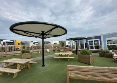 Deux abris ronds avec une toile tendue au colori gris clair, situés à l'extérieur dans un espace de détente comprenant des tables et bancs en bois.